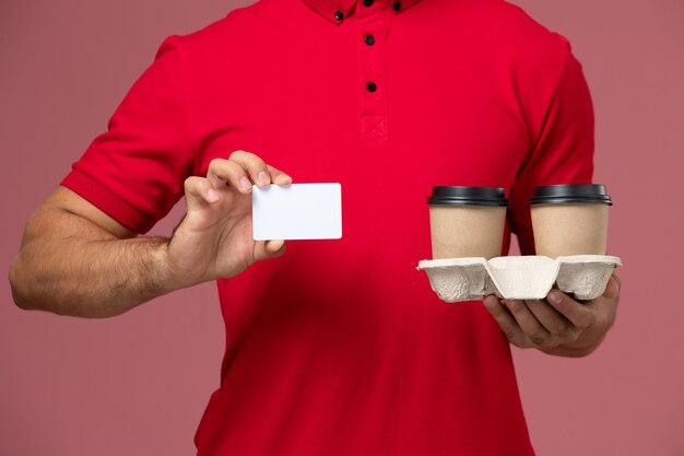 Courier masculino de vista frontal cercana en uniforme rojo sosteniendo tazas de café de entrega marrón con tarjeta blanca en la pared rosa