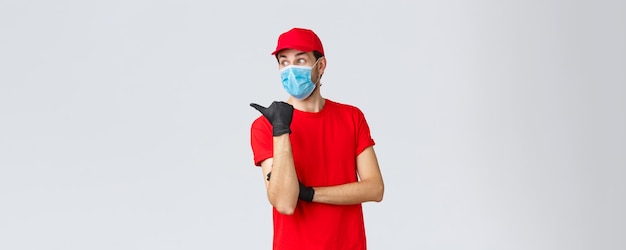 Covid-19, autocuarentena, concepto de compras y envío online. Mensajero sorprendido con uniforme rojo, mascarilla y guantes, apuntando con el pulgar y mirando a la izquierda impresionado, leyendo con curiosidad la promoción, mostrando el camino