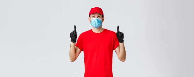 Covid-19, autocuarentena, concepto de compras y envío online. Repartidor sonriente, gorra y camiseta de uniforme rojo, máscara médica con guantes de goma, mirando hacia arriba curioso, letrero de lectura o promoción