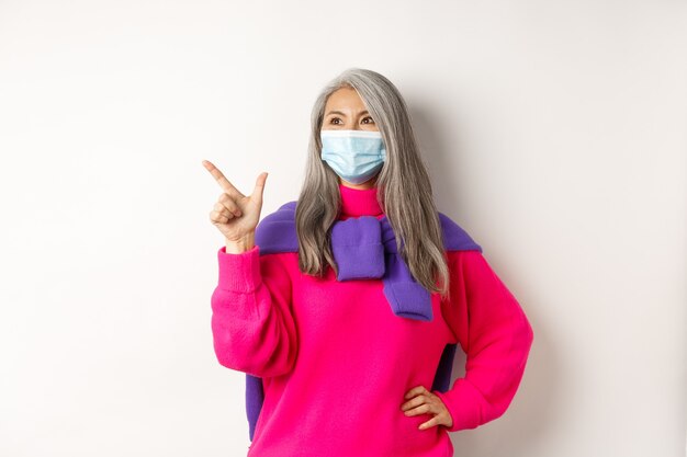 Covid-19, concepto de cuarentena y bloqueo. Elegante abuela asiática en mascarilla apuntando y mirando a la esquina superior izquierda, mostrando el logo y sonriendo, fondo blanco.