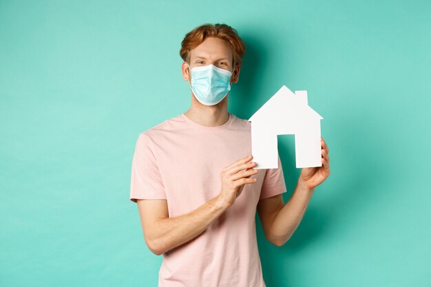 Covid-19 y concepto inmobiliario. Joven feliz en mascarilla mostrando el recorte de la casa de papel y sonriendo, ofrece propiedad en venta, de pie sobre fondo de menta.