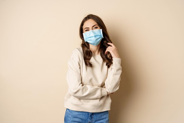 Covid-19, concepto de pandemia y cuarentena. Hermosa mujer moderna con mascarilla médica, brazos cruzados en el pecho, aspecto seguro, fondo beige.