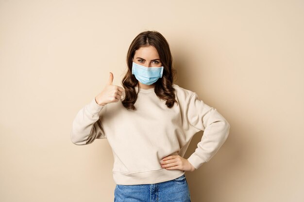 Covid-19, concepto de pandemia y cuarentena. Mujer joven usa mascarilla médica durante el brote de coronavirus omicron, mostrando los pulgares hacia arriba, de pie sobre fondo beige