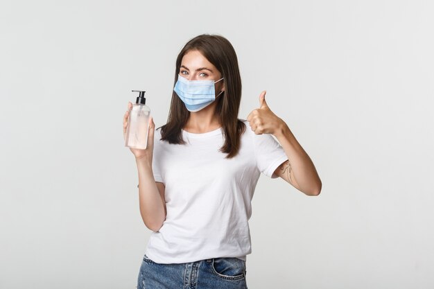 Covid-19, concepto de salud y distanciamiento social. Retrato de niña sonriente satisfecha con máscara médica, mostrando el pulgar hacia arriba y desinfectante de manos.