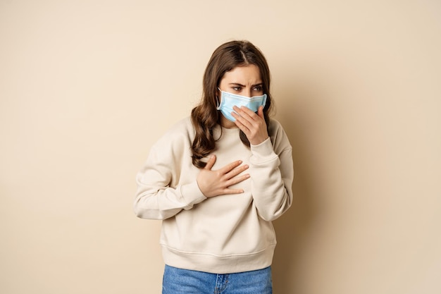 Covid-19 y concepto de salud. Mujer enferma con mascarilla médica tosiendo, sintiéndose enferma con garganta agria, de pie sobre fondo beige