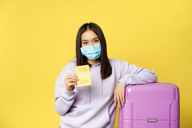 Covid-19 y concepto sanitario. Turismo. Joven turista asiática muestra su pasaporte de salud, certificado de vacunación internacional contra el coronavirus, de pie con suticase.