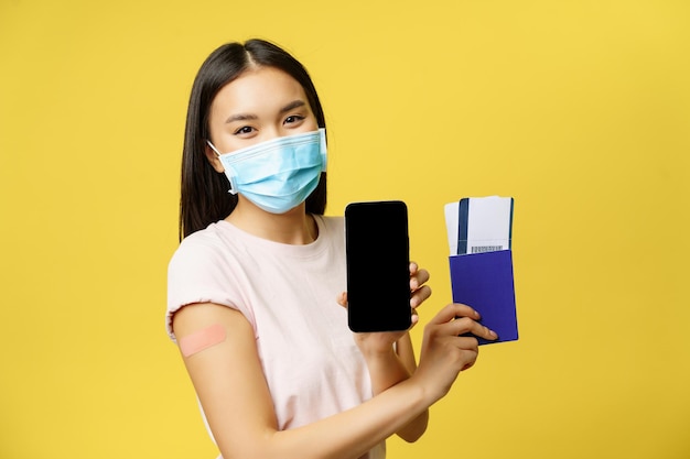 Covid-19 y concepto de turismo. Sonriente mujer asiática vacunada, mostrando la pantalla del teléfono inteligente, interfaz de la aplicación, pasaporte con boletos para vacaciones, fondo amarillo