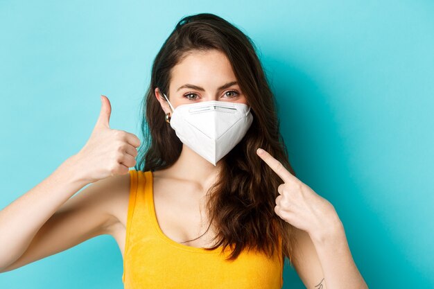 Covid-19, coronavirus y distanciamiento social. Use mascarilla. Mujer sonriente en respirador médico apuntando a la cara, mostrando el pulgar hacia arriba, de pie sobre fondo azul.