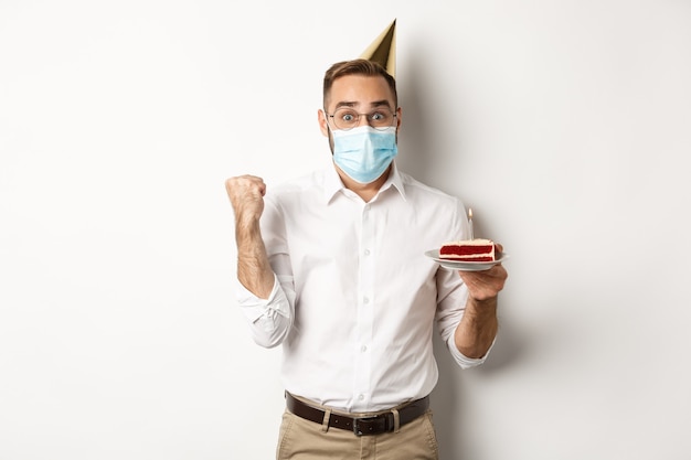 Covid-19, distanciamiento social y celebración. Hombre feliz cumpleaños esperanzado en mascarilla, sosteniendo pastel de cumpleaños y regocijo, de pie