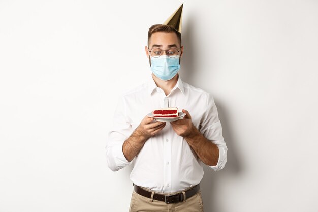 Covid-19, distanciamiento social y celebración. Hombre mirando emocionado por el pastel de cumpleaños, con máscara médica de la pandemia de coronavirus