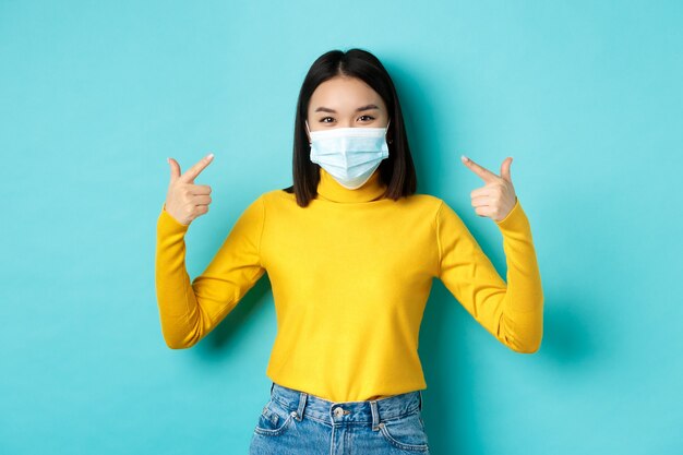 Covid-19, distanciamiento social y concepto de pandemia. Joven asiática se protege del coronavirus, señalando con el dedo su máscara médica, de pie sobre fondo azul