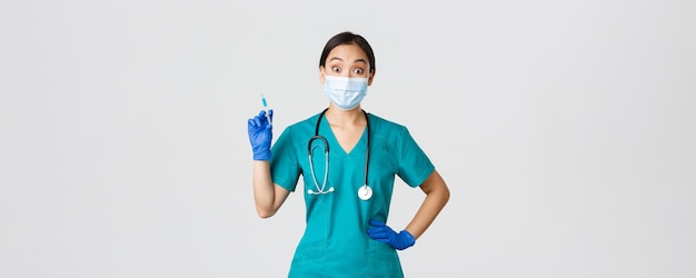 Covid-19, enfermedad por coronavirus, concepto de trabajadores de la salud. Divertida médica asiática sonriente, doctora con máscara médica y guantes de goma, jeringa preparada con vacuna para inyección, fondo blanco.