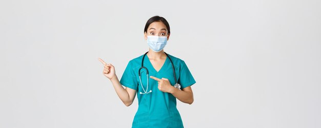Covid-19, enfermedad por coronavirus, concepto de trabajadores de la salud. Doctora asiática sorprendida e interesada, médico con máscara médica y matorrales apuntando con el dedo en la esquina superior izquierda, fondo blanco.