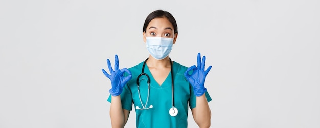 Covid-19, enfermedad por coronavirus, concepto de trabajadores de la salud. Emocionado e impresionado médico asiático, enfermera con máscara médica y guantes de goma que muestran un gesto correcto, aprueban o elogian el buen trabajo.