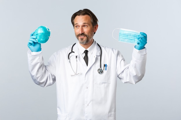 Covid-19, prevención de virus, trabajadores sanitarios y concepto de vacunación. Doctor complacido con guantes y bata blanca que muestra un respirador médico y una máscara, explica la importancia de usar PPE durante el coronavirus.
