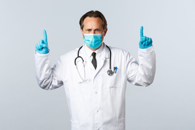 Covid-19, prevención de virus, trabajadores sanitarios y concepto de vacunación. Doctor loco serio con máscara médica y guantes apuntando con el dedo hacia arriba decepcionado, explicar las medidas de cómo luchar contra la enfermedad