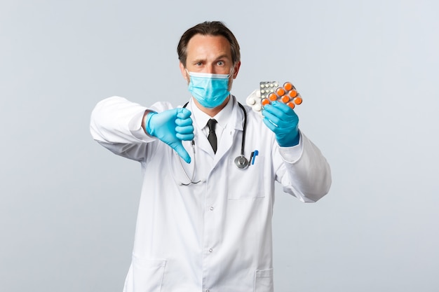 Covid-19, prevención de virus, trabajadores sanitarios y concepto de vacunación. Médico serio disgustado con máscara médica y guantes, muestra el pulgar hacia abajo y la medicación, píldoras de mala calidad, prescripción incorrecta