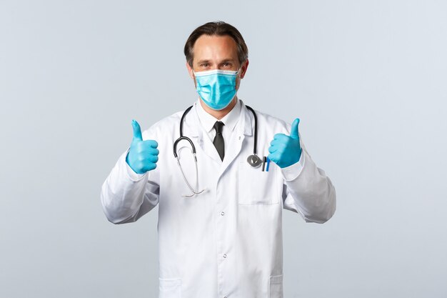 Covid-19, prevención de virus, trabajadores sanitarios y concepto de vacunación. Seguro médico guapo con máscara médica y guantes, muestra el pulgar hacia arriba, anima o recomienda los servicios de la clínica