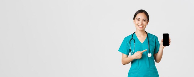 Covid-19, trabajadores de la salud y concepto de medicina en línea. Sonriente doctora asiática, médica en matorrales apuntando con el dedo a la pantalla del móvil, recomienda descargar chequeo, aplicación de consulta