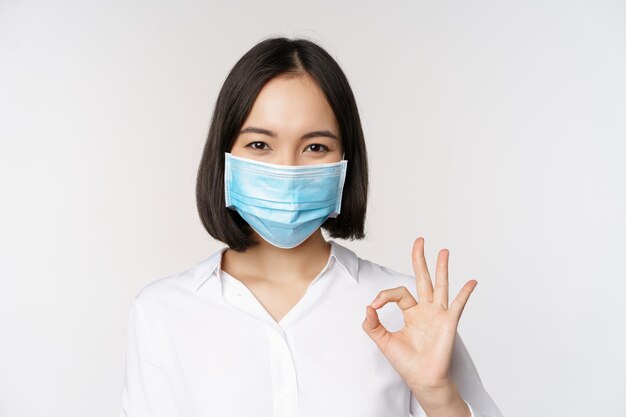 Covid y concepto de salud Retrato de una mujer asiática con mascarilla médica y mostrando un signo de estar bien de pie sobre fondo blanco.