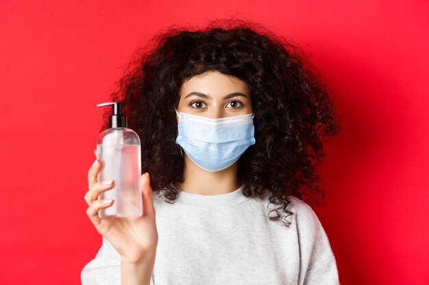 Covid pandemia y concepto de cuarentena mujer joven en máscara médica que muestra una botella de desinfectante de manos d ...