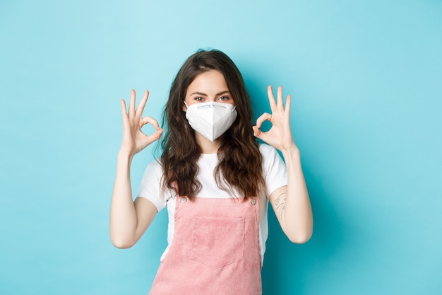 Covid, salud y concepto de pandemia. Hermosa niña satisfecha con respirador, máscara médica que muestra la aprobación de signo bien, uisng medidas de prevención del coronavirus, fondo azul.
