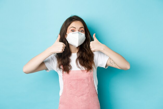 Covid, salud y concepto de pandemia. Mujer joven alegre en respirador mostrando los pulgares hacia arriba en señal de aprobación, recomendar algo, de pie contra el fondo azul.