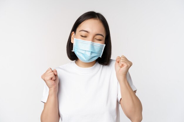 Covid19 concepto médico y de salud Mujer asiática entusiasta con mascarilla médica bailando y celebrando ganar lograr el objetivo de pie sobre fondo blanco