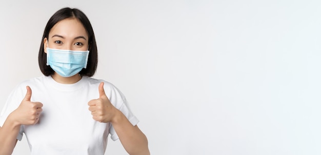 Covid19 concepto de salud y personas Joven mujer coreana feliz con máscara médica mostrando los pulgares hacia arriba de pie en camiseta sobre fondo blanco