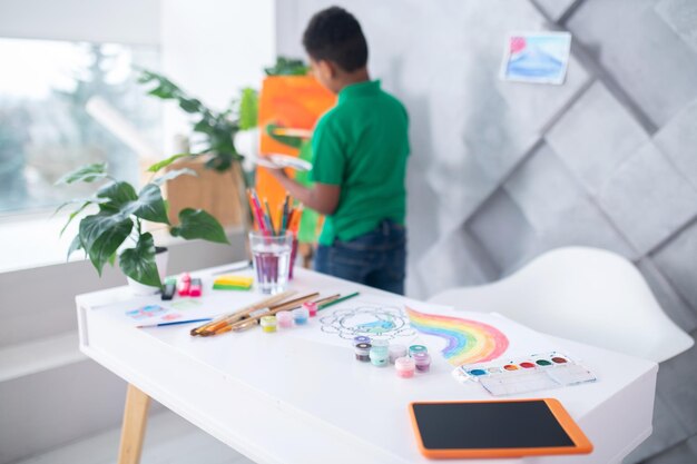 Creación. Suministros de arte y tableta en una mesa blanca y detrás de un niño en edad escolar con una camiseta verde de pie cerca del caballete de espaldas a la cámara en una habitación luminosa