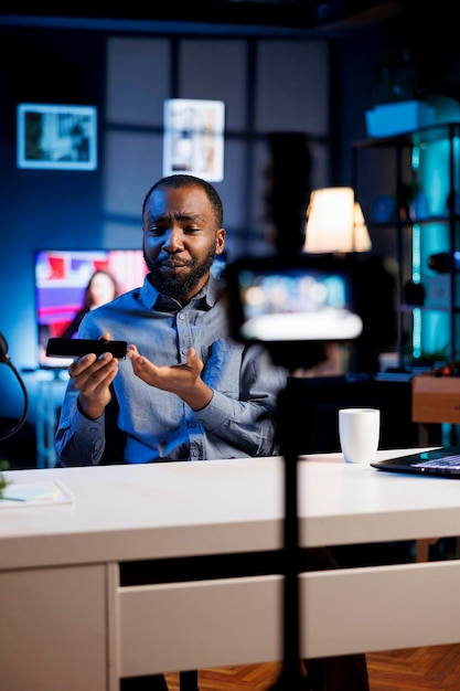 Foto gratuita creador de contenido afroamericano analizando un teléfono inteligente recién lanzado recibido de un patrocinador haciendo una revisión honesta para un canal de transmisión en línea anfitrión de un programa de internet revisando una marca de teléfono móvil