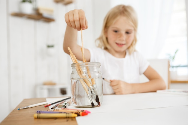 Creativa y alegre chica rubia con pecas cepillo profundo en el agua. Niña rubia pintando con un pincel. Actividades artísticas para niños.