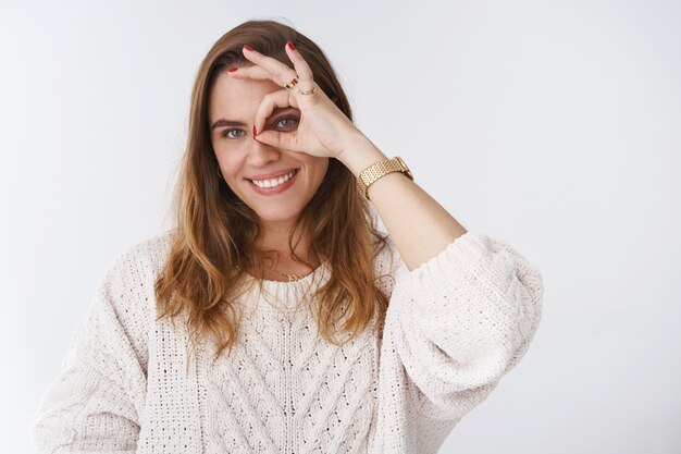 Creativa, carismática, coqueta, atractiva y encantadora mujer europea jugando contigo divirtiéndote sonriendo ampliamente dientes blancos haciendo un círculo bien bien gesto mirando a través de un curioso fondo blanco complacido