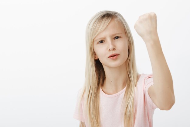 Creceré y te mostraré. Retrato de niña enojada disgustada con cabello rubio, levantando el puño cerrado y agitándolo en gesto amenazador, frunciendo el ceño, regañando a alguien por una pared gris