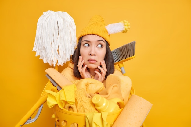 La criada asiática mantiene las manos en la cara mira hacia otro lado usa diferentes herramientas de limpieza para llevar la casa en orden posa cerca de la canasta de lavandería en amarillo vivo
