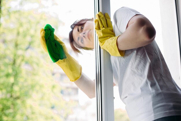 Criada con camiseta blanca y guantes protectores amarillos limpiando vidrios con un trapo Vista de ama de casa morena limpiando una gran ventana sucia Concepto de tareas domésticas y servicio de apartamentos