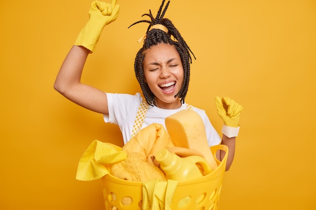 La criada de la mujer feliz baila despreocupada mantiene poses de brazos levantados cerca del lavabo se divierte mientras hace las tareas del hogar canta una canción aislada sobre un fondo amarillo vivo. Concepto de limpieza