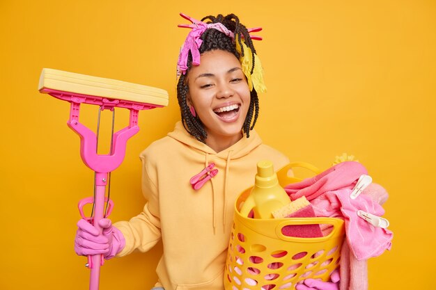 La criada de piel oscura alegre sonríe ampliamente usa sudadera y guantes de goma protectores sostiene la canasta de lavandería y la fregona feliz de terminar las tareas del hogar aislado en amarillo