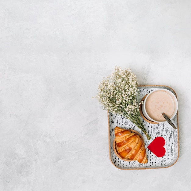 Croissant, adorno de corazón, plantas y taza de bebida en bandeja.