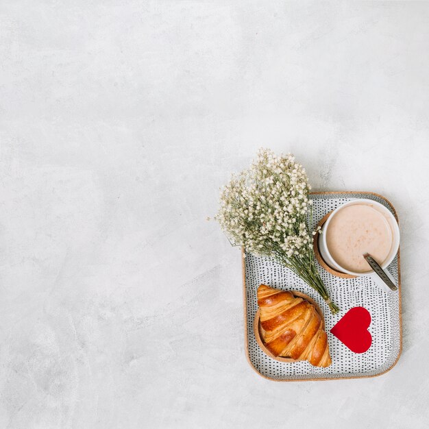 Croissant, adorno de corazón, plantas y taza de bebida en bandeja.