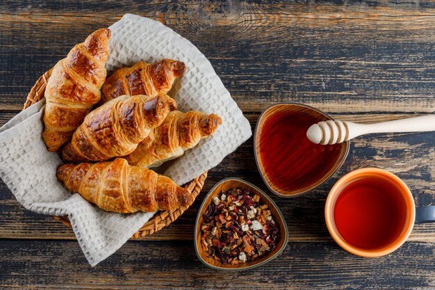 Croissant en una canasta con miel, cazo, té, hierbas secas en una mesa de madera