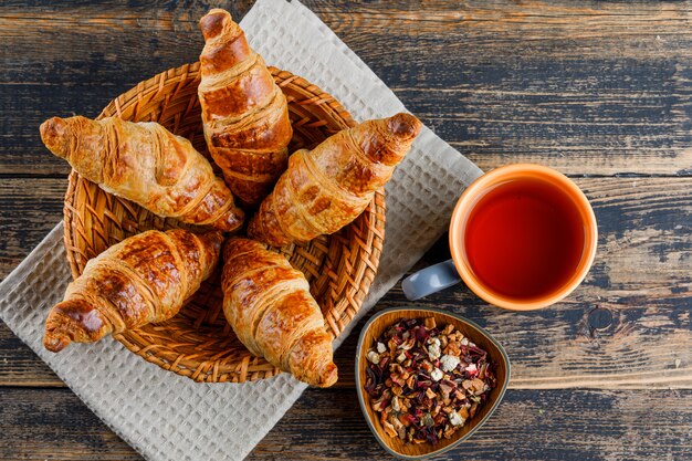 Croissant en una cesta con una taza de té, hierbas secas planas sobre una toalla de madera y cocina