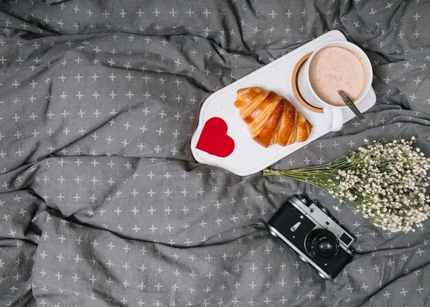 Croissant, corazón de adorno y taza de bebida en una tabla de cortar cerca de las plantas y la cámara