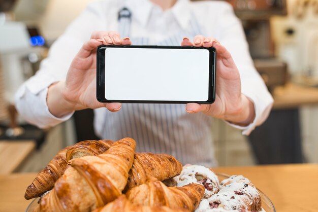 Croissant fresco delante de la mujer que sostiene el teléfono inteligente con pantalla en blanco en blanco