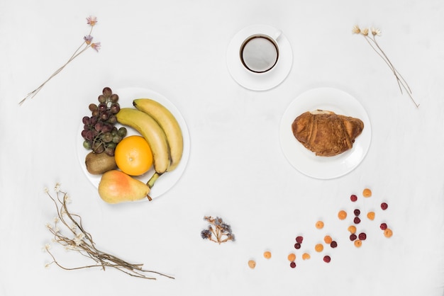 Croissant y frutas en un plato blanco con café y flores secas en el fondo blanco