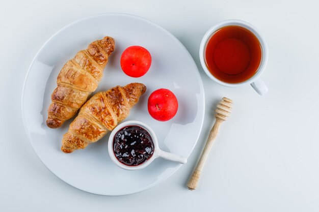 Croissant con mermelada, ciruelas, cazo, té en un plato, plano.