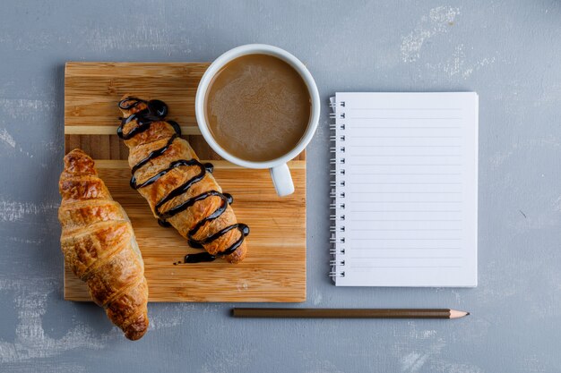 Croissant con salsa, café, cuaderno, lápiz sobre yeso y tabla de madera, plano.