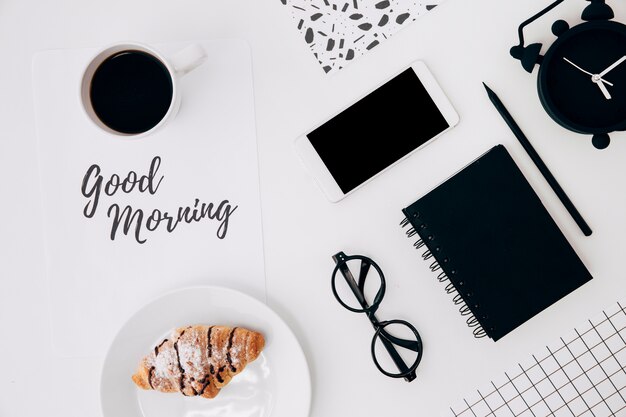 Croissant y taza de café con mensaje de buenos días en papel y artículos de oficina en escritorio blanco