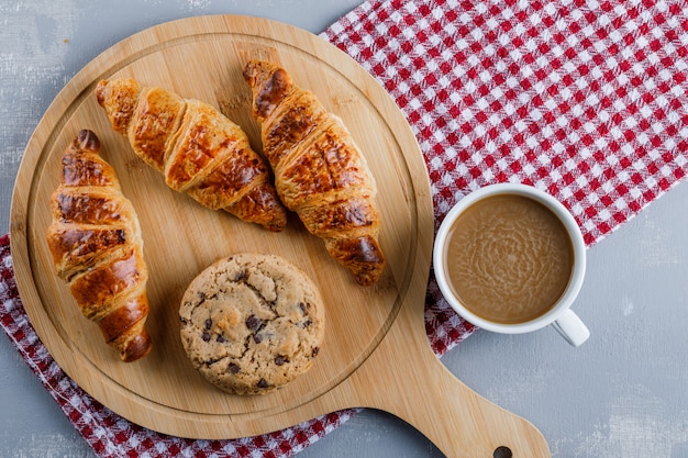 Croissants con café, galletas, tabla de cortar plana sobre yeso y tela de picnic