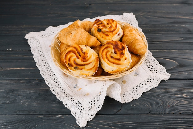 Croissants y cupcake en la cesta sobre la servilleta de encaje sobre el fondo de madera